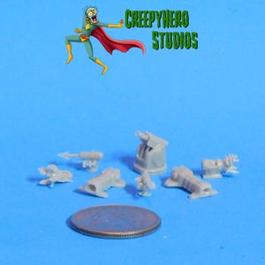 May include: A collection of small, gray plastic model pieces, including cannons and other military-themed components, are arranged on a blue surface. The image includes the text "CREEPYHERO STUDIOS" and a cartoon character.