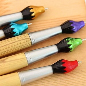 May include: A collection of artist-themed pens. The pens have a silver barrel, wooden handle, and a brush-shaped tip in various colors, including gold, blue, purple, green, and red. The pens are arranged on a light brown surface.