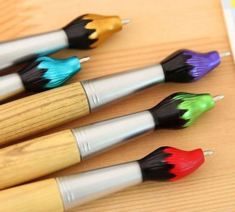 May include: A collection of artist-themed pens. The pens have a silver barrel, wooden handle, and a brush-shaped tip in various colors, including gold, blue, purple, green, and red. The pens are arranged on a light brown surface.