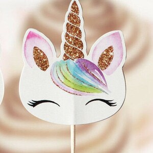 May include: A white unicorn cupcake topper with a gold glitter horn and rainbow mane. The unicorn has closed eyes and a small smile.