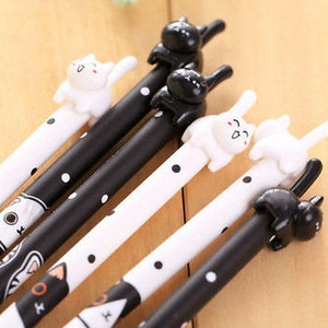 May include: A collection of five pens featuring black and white polka dot patterns. Each pen is topped with a small, three-dimensional cat figure in either solid black or white. The barrels display stylized cat face illustrations at the base.
