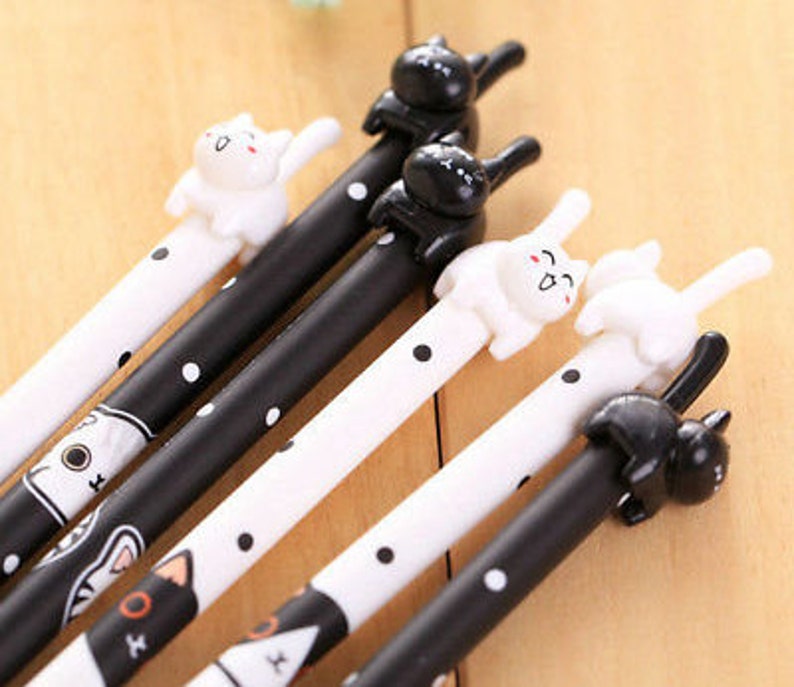 May include: A collection of five pens featuring black and white polka dot patterns. Each pen is topped with a small, three-dimensional cat figure in either solid black or white. The barrels display stylized cat face illustrations at the base.