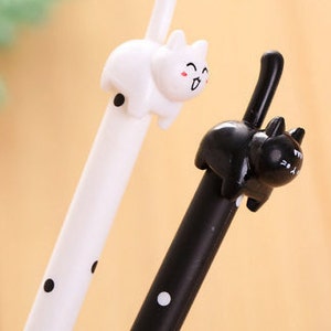 May include: Two gel pens featuring cat-shaped toppers. One pen is white with black polka dots and a white cat topper with a smiling face. The other pen is black with white polka dots and a black cat topper with a smiling face and an upright tail.