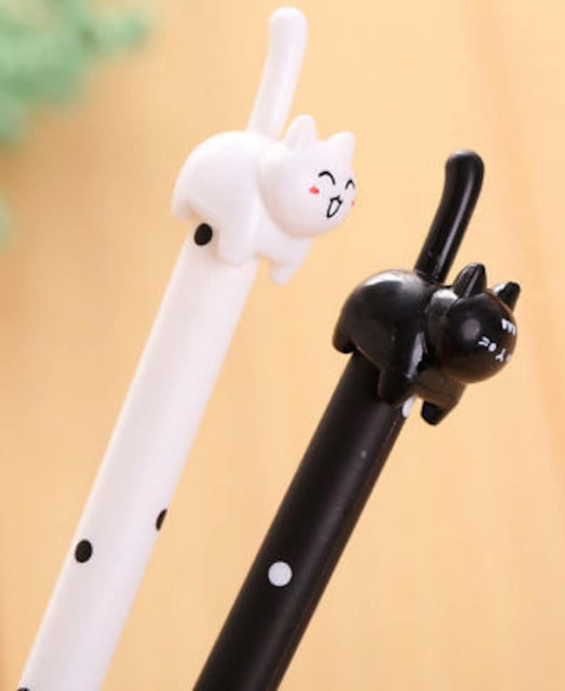 May include: Two gel pens featuring cat-shaped toppers. One pen is white with black polka dots and a white cat topper with a smiling face. The other pen is black with white polka dots and a black cat topper with a smiling face and an upright tail.