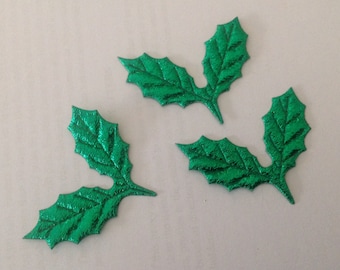May include: Three bright green holly leaf embellishments with textured surfaces. These decorative leaves are ideal for holiday crafts and add a festive touch to any project. The leaves are arranged on a neutral background.