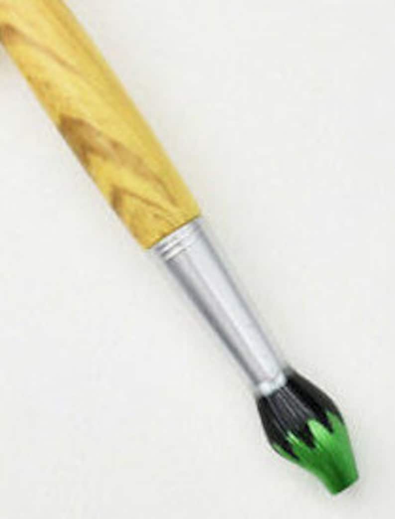 May include: A paintbrush with a light brown wooden handle and a silver ferrule. The bristles are black with green tips. The brush is angled diagonally against a white background.