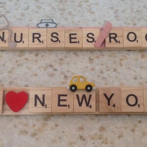 Könnte beinhalten: Zwei Reihen mit hölzernen Buchstabenkacheln, die "NURSES ROCK" und "I LOVE NEW YORK" buchstabieren, mit kleinen Dekoartikeln auf jeder Kachel. Die erste Reihe hat ein Stethoskop, eine Krankenschwestermütze und ein Pflaster. Die zweite Reihe hat eine Miniatur der Freiheitsstatue, ein rotes Herz, ein gelbes Spielzeugauto und ein Stück Pizza.