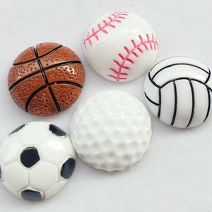May include: Five small, round, sports-themed decorations. A white baseball with red stitching, a brown basketball, a white golf ball with dimples, a white volleyball with black lines, and a white soccer ball with black panels.