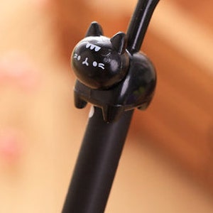 May include: A black ink pen featuring a three-dimensional black cat figure attached to the top. The cat has a smooth, glossy finish with white painted details on its face, including eyes, whiskers, and a small mouth, and a long upright tail.