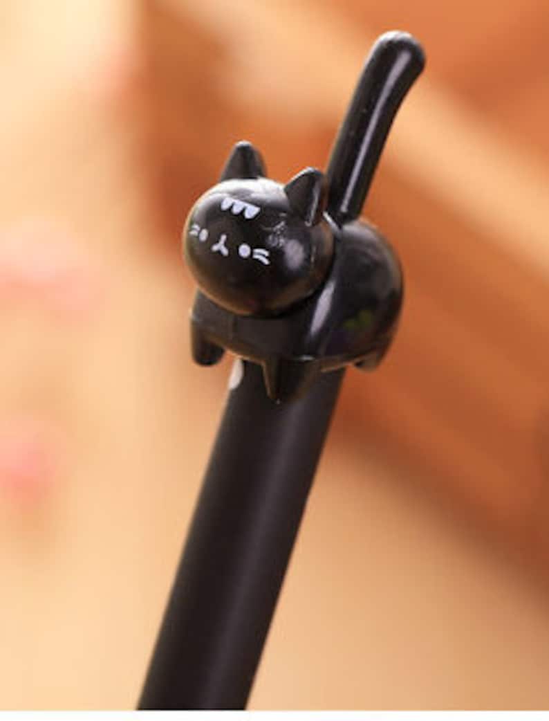 May include: A black ink pen featuring a three-dimensional black cat figure attached to the top. The cat has a smooth, glossy finish with white painted details on its face, including eyes, whiskers, and a small mouth, and a long upright tail.