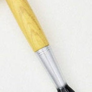 May include: A pen designed to resemble a paintbrush. It features a light brown wooden handle, a silver metal ferrule, and a purple and black brush tip. The pen is angled diagonally against a white background.