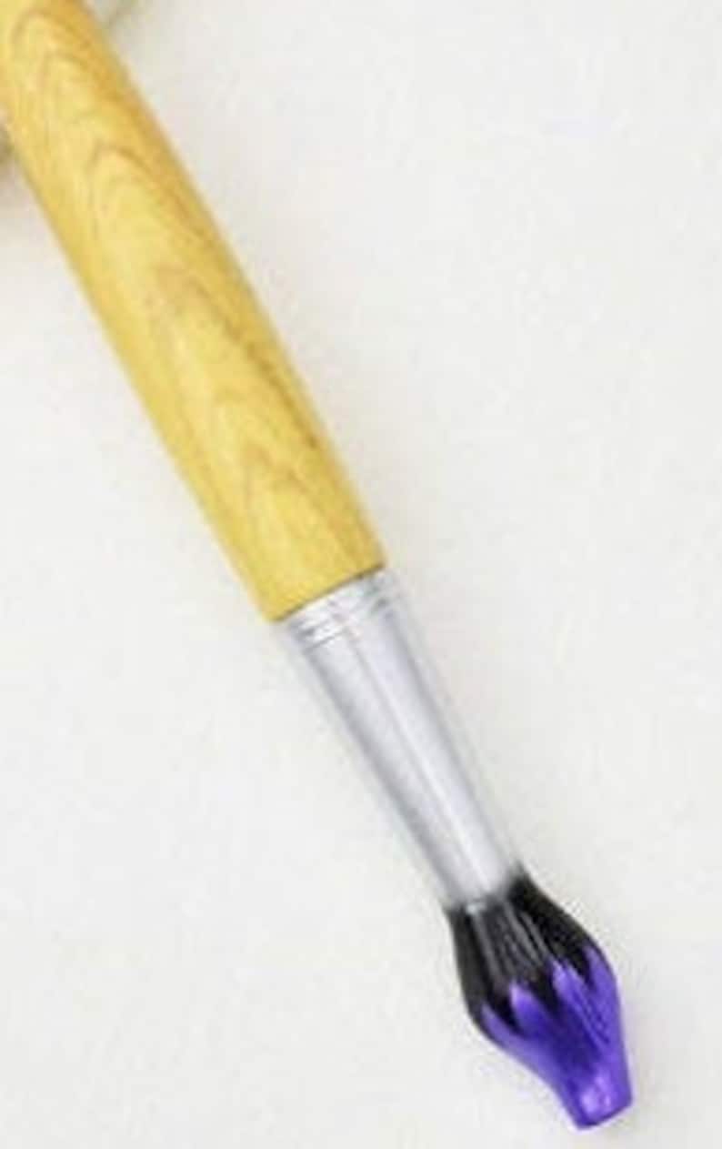 May include: A pen designed to resemble a paintbrush. It features a light brown wooden handle, a silver metal ferrule, and a purple and black brush tip. The pen is angled diagonally against a white background.