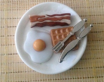 Può includere: Un piatto bianco con un uovo fritto, una fetta di pancetta, un waffle e due posate d'argento.