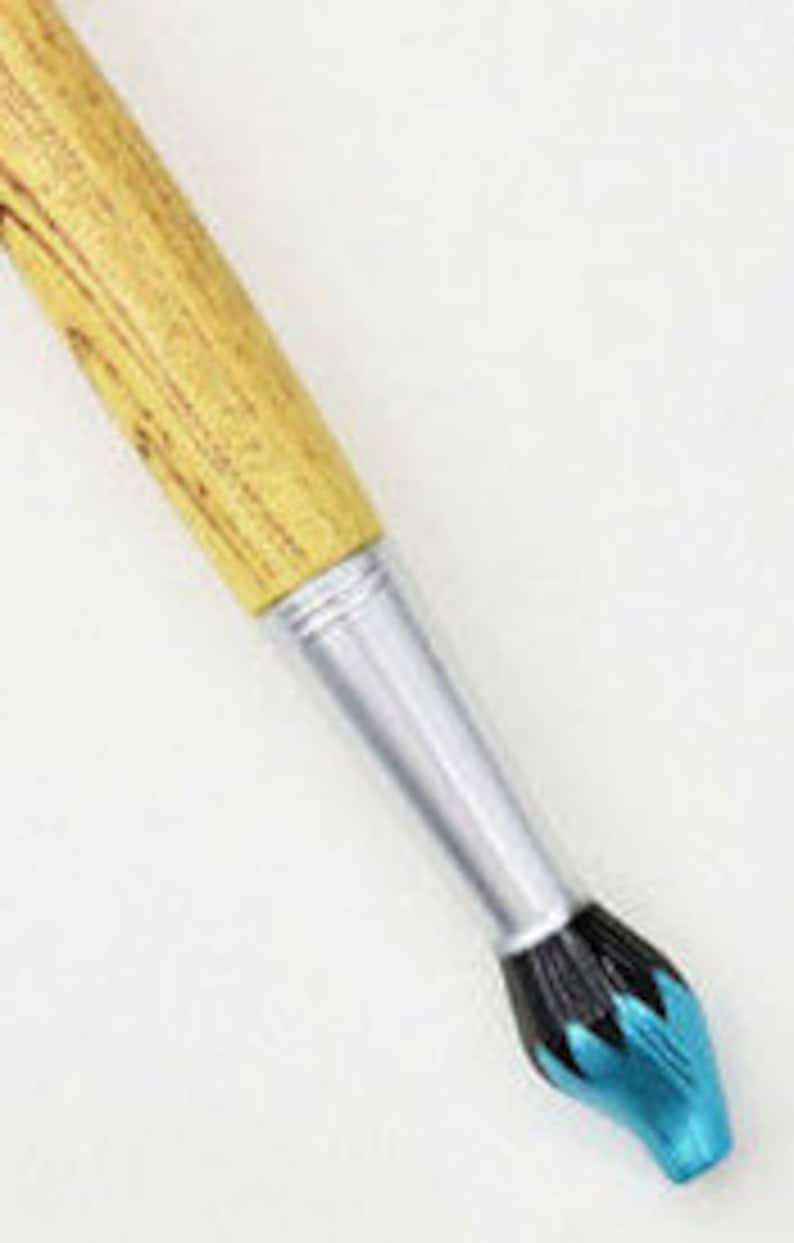 May include: Close-up of a paintbrush with a wooden handle and a silver ferrule. The bristles are black and turquoise. The brush is angled diagonally against a white background.