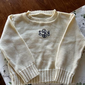 May include: A cream-colored knitted sweater with a black embroidered monogram "S3G" on the front.