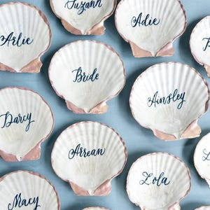 May include: White scallop shell place cards with pink edges and names written in dark blue script. The word "Bride" is visible on one shell. Other names include Arlie, Darcy, Ainsley, and Lola. The shells are arranged on a light blue surface.