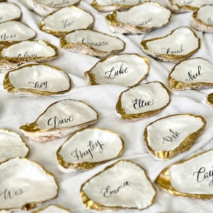 May include: White oyster shells with gold trim, used as place cards for a wedding or party. Each shell has a name written in black calligraphy. Names include: Gess, Alam, Yac, Vanessa, Wes, Troy, Dave, Hayley, Emma, Frank, Luke, Neil, Ellie, Rush, and Cau.