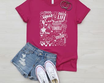 May include: A magenta t-shirt with a white graphic design featuring the words "Squad," "Cheer Life," and "Tumble." The shirt is paired with denim shorts and white sneakers. A set of bracelets rests on the shirt.