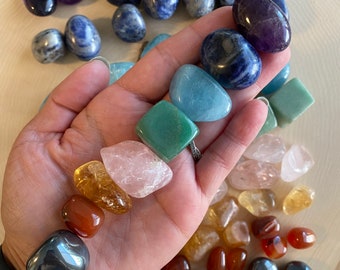May include: A selection of polished gemstones in shades of purple, blue, green, yellow, orange, pink, and grey. The stones are displayed on a wooden surface, with some held in a hand. The gemstones are smooth and rounded.