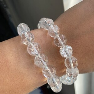 May include: Two clear quartz crystal bead bracelets. Each bracelet features large, round, crackled beads strung on a clear elastic cord. The bracelets are worn on a wrist, showcasing the translucent quality of the quartz.