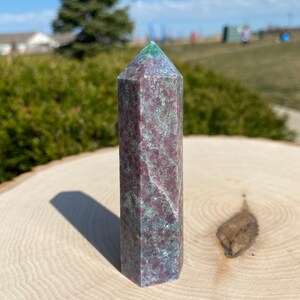 May include: A faceted crystal obelisk in shades of burgundy, green, and grey. The crystal has a flat top and stands on a light-coloured wooden surface. The background shows a natural outdoor scene.