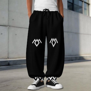 May include: Black sweatpants featuring a white geometric mountain-like logo on each leg. The pants have a white elastic waistband with a black drawstring and black and white chevron-patterned cuffs at the ankles, paired with black and white sneakers.