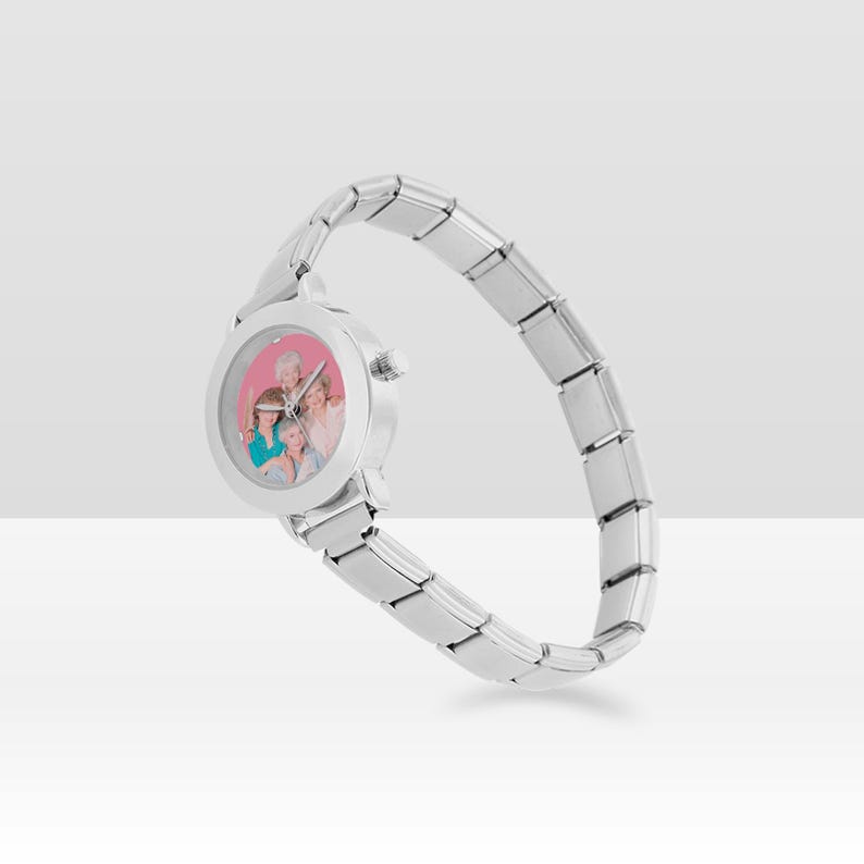 May include: A silver wristwatch with a round face displaying a group photograph. The watch features a flexible strap made of rectangular links. The watch face has a pink background.