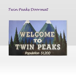 Può includere: Zerbino rettangolare con la scritta "WELCOME TO TWIN PEAKS" e una scena di montagna e foresta. Lo zerbino include anche la popolazione di 51.200. Il testo "Twin Peaks Doormat" si trova nell'angolo in alto a sinistra.