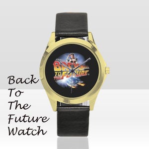 May include: A gold-toned watch with a black leather band. The watch face features the "Back to the Future" movie logo and a scene from the film. The words "Back To The Future Watch" are written on the left side.