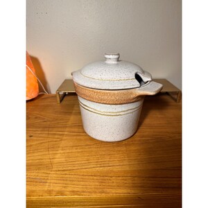 May include: A ceramic lidded serving dish with a spoon rest. The dish is speckled white with a brown band and gold stripes. The lid has a small handle. The dish is on a wooden surface.