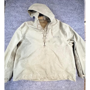 May include: A vintage olive drab US Navy pullover anorak with a hood and lace-up neckline. The anorak features the text "U.S.N." on the front. The sleeves have buttoned cuffs. The anorak is made of a durable, possibly water-resistant material.