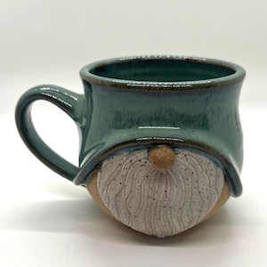 Peut inclure: Une tasse en céramique avec une partie supérieure émaillée bleu sarcelle formant un bord de chapeau au-dessus d'une barbe blanche texturée et tachetée et d'un petit nez brun. La base est en argile beige non émaillée avec une anse incurvée.
