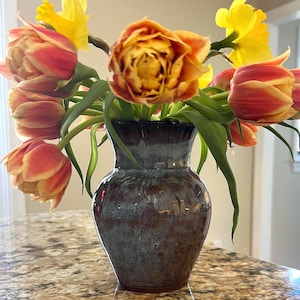 May include: A ceramic vase with a dark brown and blue glaze holds a bouquet of flowers. The bouquet includes yellow daffodils and orange and red tulips. The vase is sitting on a granite countertop.