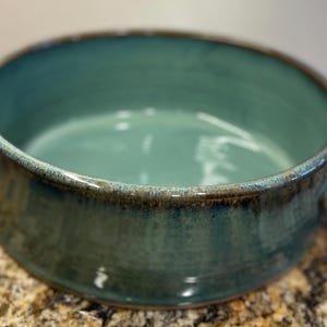 May include: A teal ceramic bowl with a rounded shape and two small handles. The bowl has a glossy finish with a darker rim and a speckled, textured appearance. The bowl is likely handmade.
