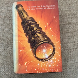 May include: A book cover featuring a detailed illustration of a brass telescope with rope detailing, set against a fiery orange and yellow background with sparkling light effects. Text reads: "War in heaven - and the fate of the worlds lies in the balance. It's all up to will and Lyra now..."