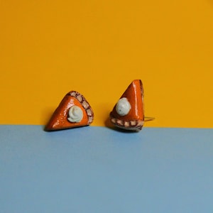 May include: A pair of small, handmade pumpkin pie earrings. The earrings are orange with a white swirl of whipped cream on top. They are on a blue and yellow background.