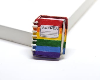 May include: A small rainbow-colored notebook with a white spiral binding. The cover has the word "AGENDA" printed on it.