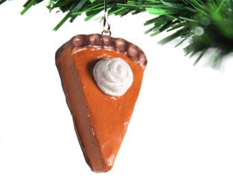 Peut inclure: Une décoration de Noël en forme de tarte à la citrouille de couleur marron et orange avec une touche de crème fouettée blanche sur le dessus. La décoration est suspendue à un sapin de Noël artificiel vert.