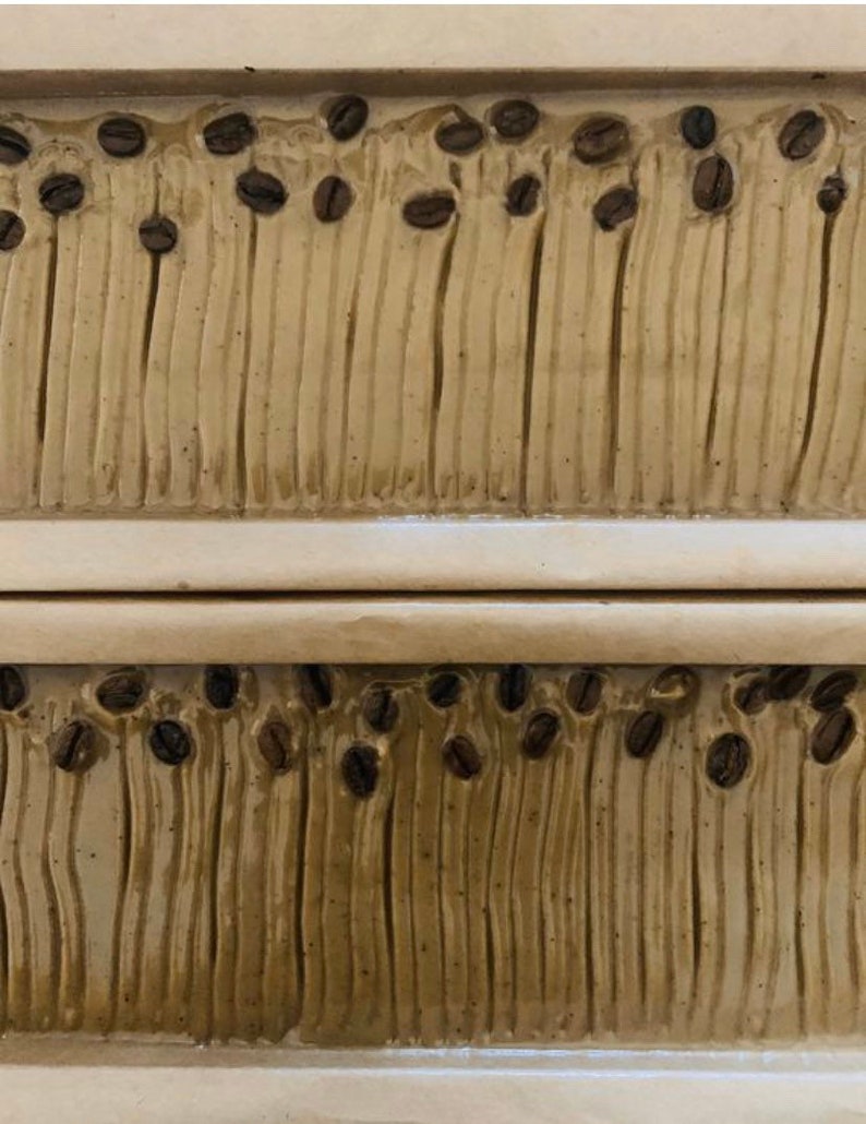 May include: Two rectangular bars of soap with a brown and beige swirl pattern. The soap is decorated with small brown coffee beans.