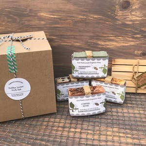 May include: A gift set for coffee lovers featuring four bars of handmade organic soap in various coffee-inspired scents. The soaps are wrapped in brown paper and tied with twine. The set also includes a wooden soap dish and a brown cardboard box with a label that reads "Coffee Lover Gift Set".