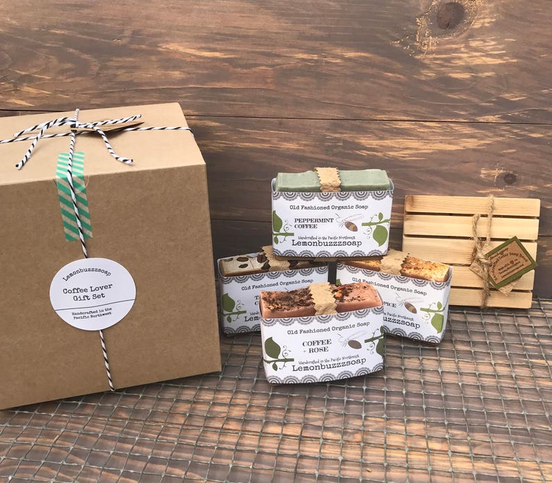 May include: A gift set for coffee lovers featuring four bars of handmade organic soap in various coffee-inspired scents. The soaps are wrapped in brown paper and tied with twine. The set also includes a wooden soap dish and a brown cardboard box with a label that reads "Coffee Lover Gift Set".