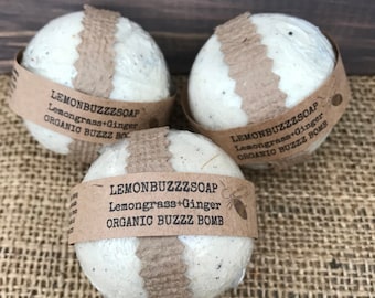 Puede incluir: Tres bombas de baño redondas y blancas con bandas de papel marrón. Cada banda tiene el texto "LEMONBUZZZSOAP Lemongrass+Ginger ORGANIC BUZZZ BOMB". Las bombas están sobre una superficie de arpillera.