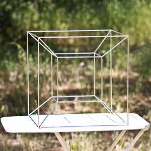 May include: A silver wire cube sculpture, standing on a white table, against a blurred background of green grass and trees.