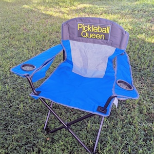 Puede incluir: Silla plegable azul y gris con el texto "Pickleball Queen" en el respaldo. La silla tiene respaldo de malla, reposabrazos con portavasos y estructura de metal negro. Se encuentra sobre una superficie de césped.