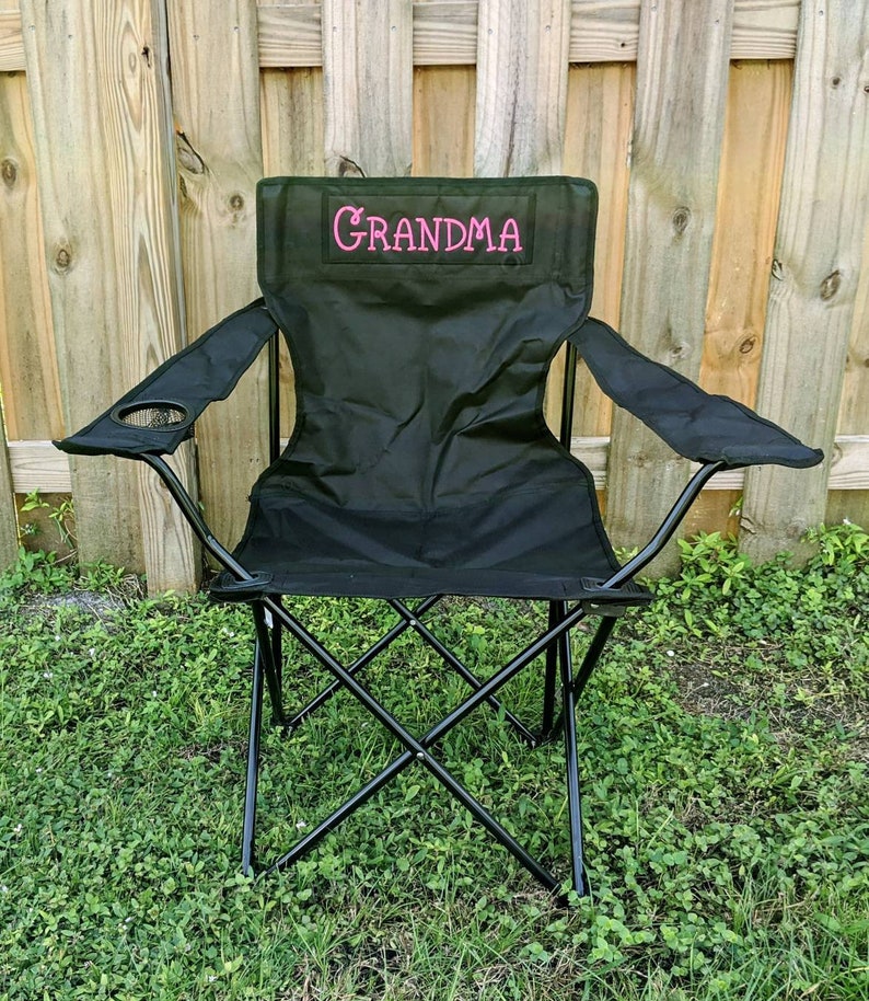 Puede incluir: Silla plegable negra con un portavasos y un "GRANDMA" bordado en rosa en el respaldo.