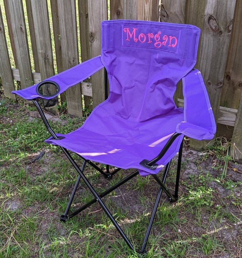 Puede incluir: Silla plegable de color morado con marco negro y portavasos. La silla tiene el nombre "Morgan" bordado en la parte posterior en color rosa.