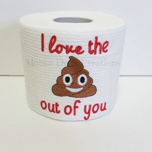 Op de afbeelding: Een witte toiletrol met rode borduurwerk dat "I love the out of you" zegt met een bruine poep emoji met een smiley in het midden.