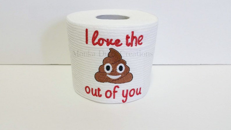 Op de afbeelding: Een witte toiletrol met rode borduurwerk dat "I love the out of you" zegt met een bruine poep emoji met een smiley in het midden.
