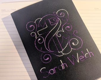 Op de afbeelding: Zwart notitieboek met een geborduurd ontwerp in wit en paars. Het ontwerp bevat een gestileerde letter "S" en andere decoratieve elementen. De naam "Sarah Welch" is eronder in paars geborduurd.