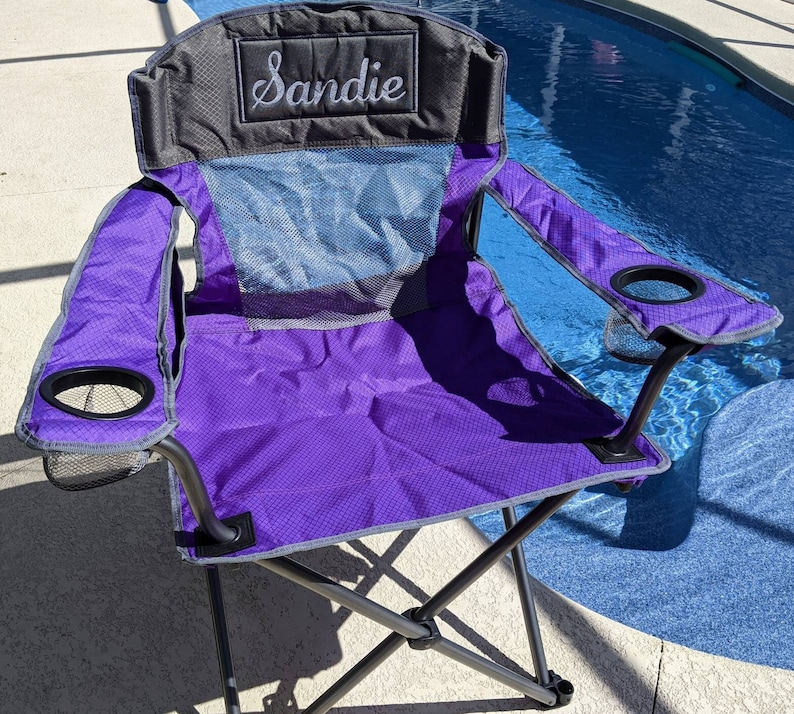 May include: Purple folding chair with a black frame and a gray mesh back. The chair has two cup holders and a personalized name tag that says "Sandie".